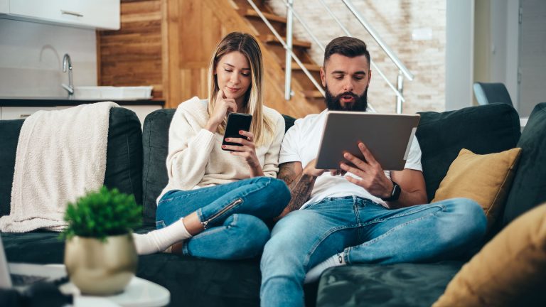 Zwei Personen sitzen auf der Couch und haben jeweils ein iPhone und ein Tablet vor sich in der Hand.