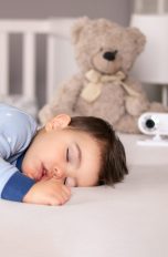 Ein Kind liegt schlafend im Bett, im Hintergrund ein Teddy und ein Babyphone.