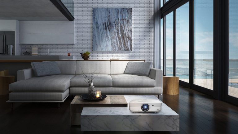 Optoma UHZ65LV Ein Beamer steht auf einem marmorierten Tisch vor einer Wohnlandschaft links neben einer bodentiefen Fensterfront.