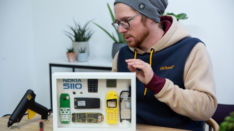 Eine Person steht hinter einem selbstgebauten Technik-Bilderrahmen. Darin befindet sich ein in seine Einzelteile zerlegtes Nokia 5110.