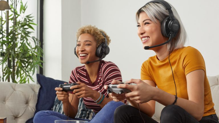 Zwei Personen sitzen auf dem Sofa, spielen auf einer PS4 und tragen dabei das Headset JBL Quantum.