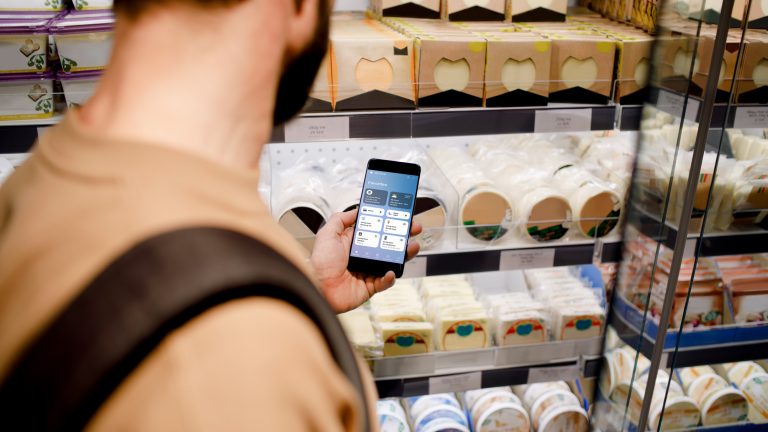 Smarte Haushaltsgeräte Eine Person steht vor einem Kühlregal und schaut auf ihr Smartphone, auf dem die Samsung SmartThings App geöffnet ist.