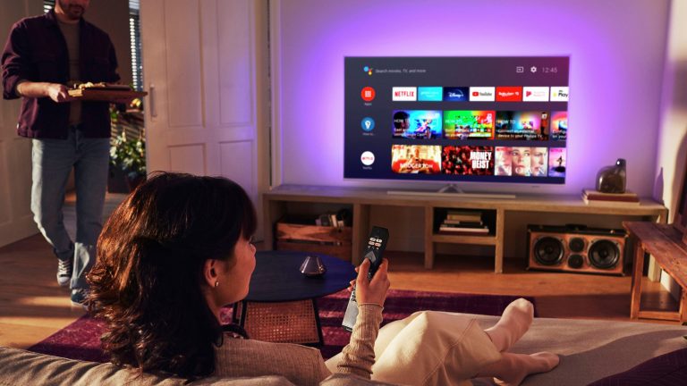 Ambilight nachrüsten Eine Person sitzt auf dem Sofa, vor ihr ist ein Philips 58PUS8506 mit angeschaltetem Ambilight zu sehen.