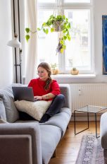Eine Frau sitzt mit ihrem Laptop auf dem Sofa in ihrem Wohnzimmer.
