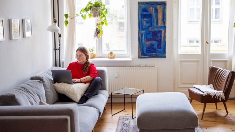 Eine Frau sitzt mit ihrem Laptop auf dem Sofa in ihrem Wohnzimmer.