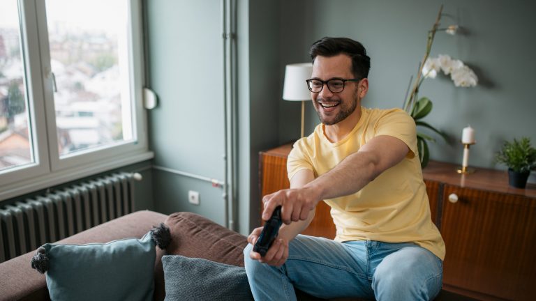 DSL, TV-Kabel & Glasfaser Ein junger Mann sitzt auf der Lehne seines Sofas, in der Hand hält er den Controller seiner Spielekonsole.