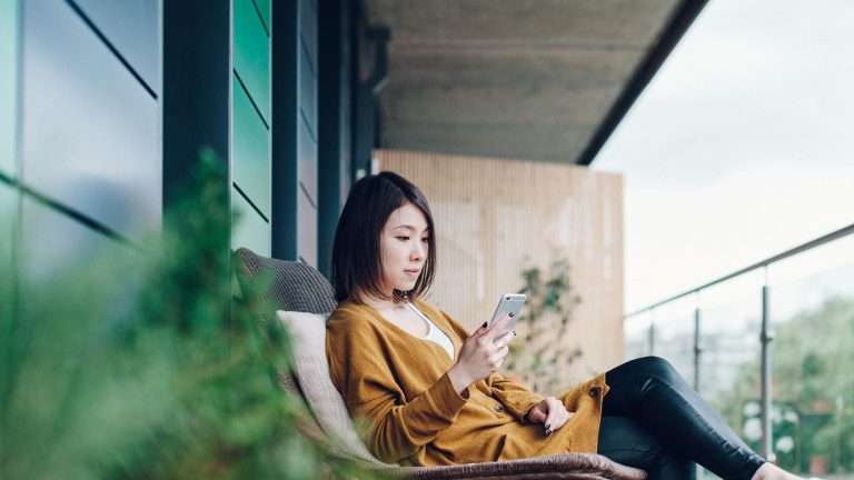 Eine Person sitzt in einem Stuhl auf einem Balkon und schaut auf ihr iPhone.