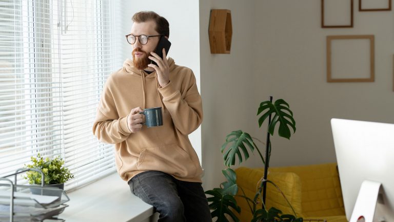 iPhone-Rufnummer unterdrücken Eine Person lehnt an einer Fensterbank. In der einen Hand hält sie einen Kaffeebecher, während sie mit der anderen telefoniert.