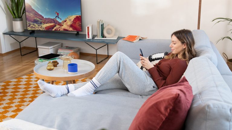 Smart TV Eine Person liegt auf einem Sofa, neben ihr sind auf einer TV-Bank ein Fernseher und eine FRITZBox_6690_Cable zu sehen.