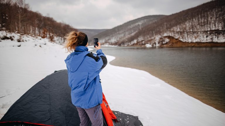 Camping-Gadgets Eine Person steht vor einem Zelt im Schnee in der Nähe eines Flusses. In der Hand hält sie ein Smartphone, mit dem sie die Landschaft fotografiert.