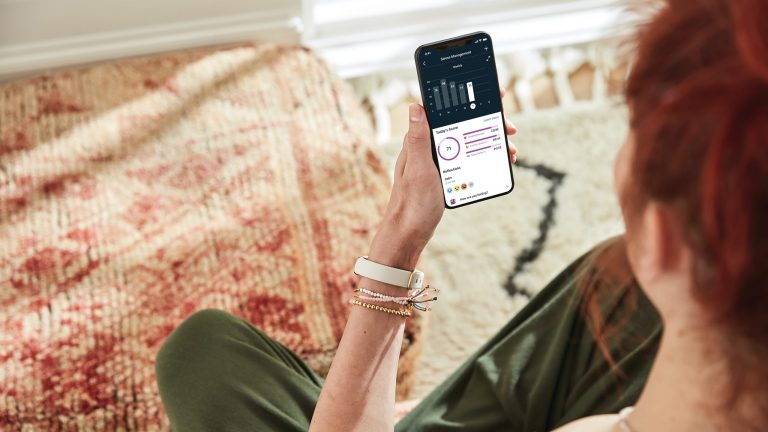 Fitbit Luxe Eine Person sitzt auf einem Teppich und schaut auf das Smartphone in ihrer Hand. Darauf wird gerade eine Statistik in der Fitbit-App angezeigt.