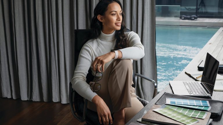 Fitbit Sense Eine Person sitzt auf einem Stuhl am Schreibtisch, ein Bein angezogen, vor ihr steht ein Laptop.