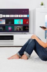 Eine Person sitzt auf einem Teppich vor einem Sofa und schaut zum Fernseher an der Wand, in der Hand eine Fernbedienung.