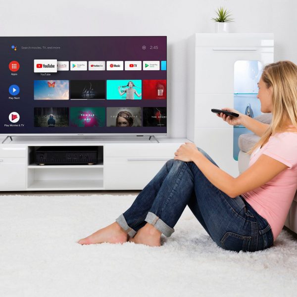 Eine Person sitzt auf einem Teppich vor einem Sofa und schaut zum Fernseher an der Wand, in der Hand eine Fernbedienung.