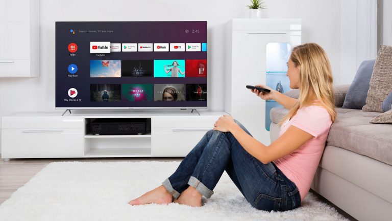 Eine Person sitzt auf einem Teppich vor einem Sofa und schaut zum Fernseher an der Wand, in der Hand eine Fernbedienung.
