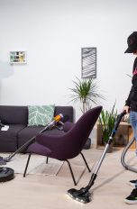 Zwei Personen saugen einen Raum mit Handstaubsauger und Bodenstaubsauger, auf dem Boden steht ein Saugroboter, an einem Sofa lehnt ein Stielsauger.