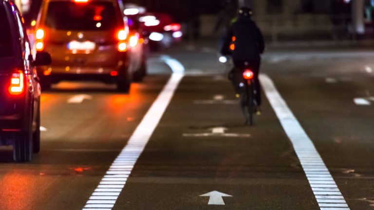 Smartes Fahrrad Auf einer Straße im Stadtverkehr steht eine Schlange von Autos mit leuchtendem Bremslicht. Daneben befindet sich ein Fahrradweg, auf dem eine Person mit einem Fahrrad fährt.