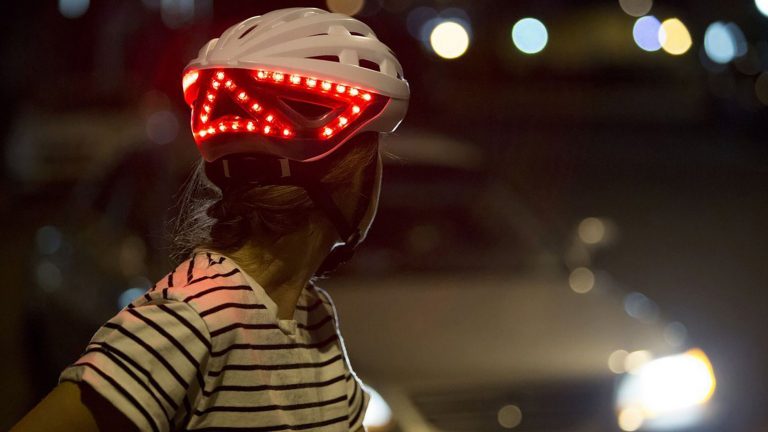 Lumos Kickstart Refreshed Eine Person trägt den Fahrradhelm Kickstart von Lumos. Sie wirft einen Blick über ihre Schulter, sodass die leuchtenden LEDs an der Rückseite des Helms zu sehen sind.