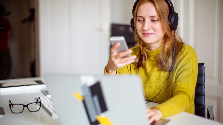 Eine Person sitzt vor einem Laptop und schaut auf ein Smartphone in ihrer Hand.