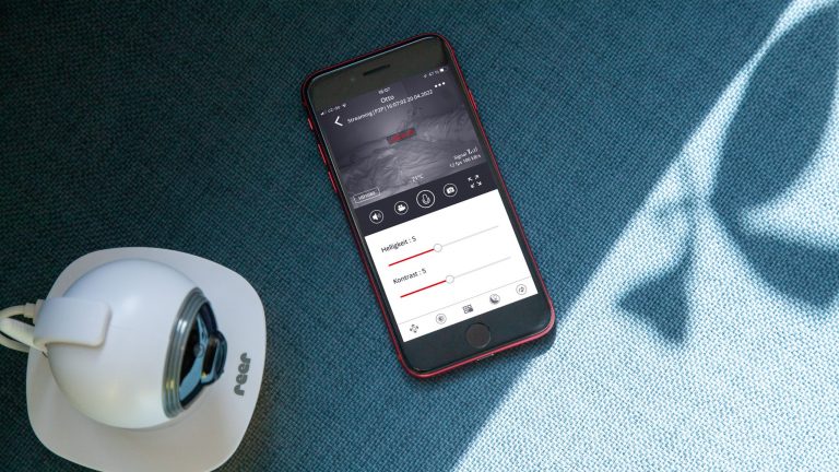 Reer Video-Babyphone IP BabyCam Smart Eine Reer IP Babycam Move steht neben einem Smartphone auf einem Sofa. Auf dem Display sind diverse Einstellungsoptionen der Kamera zu erkennen.