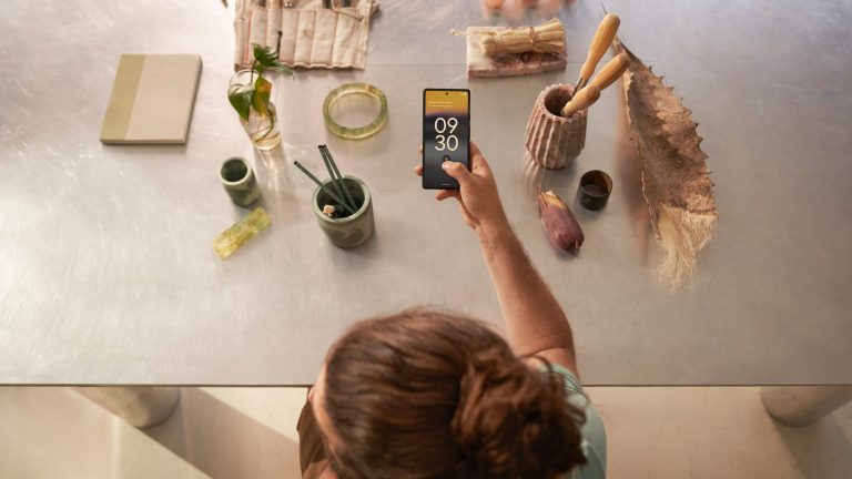 Google Pixel 6a Eine Person in Arbeitsschürze sitzt an einem Arbeitsplatz, auf dem einige Utensilien verteilt sind. In der Hand hält die Person ein Google Pixel 6a, auf dem die Uhrzeit zu erkennen ist.