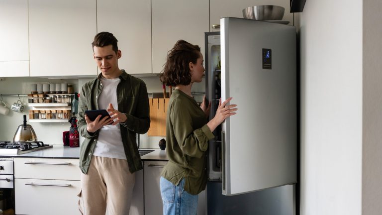 Ein Paar steht in der Küche, eine Person schaut auf ein Tablet, das sie in der Hand hält, die andere schaut in den geöffneten Kühlschrank.