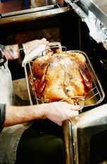 Eine Person kniet vor einem Backofen und holt ein Backhähnchen heraus.