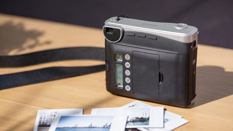 Fujifilm Instax mini 90 Neo Classic Eine Instax mini 90 Neo Classic von hinten, auf der mehrere Tasten zu sehen sind. Vor der Kamera liegen einige bereits ausgedruckte Fotos.