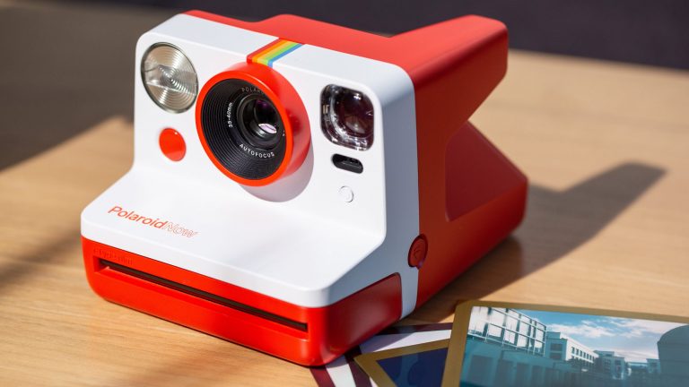 Polaroid Now Eine Polaroid-Kamera liegt auf einem Tisch, daneben mehrere Ausdrucke.