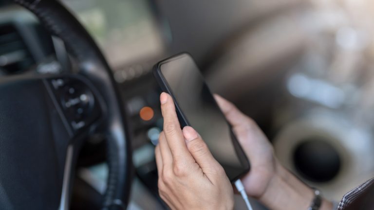 Autoradios mit Android Auto oder Apple Carplay Eine Person sitzt auf dem Gfahrersitz eines Autos und hät ein Smartphone in den Händen, dass per USB-Kabel mit dem Autoradio verbunden ist.