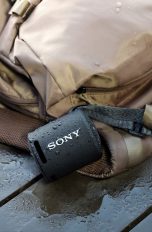 Ein schwarzer Sony SRS-XB13 hängt an einem nassen Rucksack.