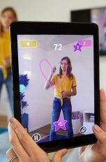 Eine Person hält ein Tablet in die Luft. Darauf ist eine junge Person zu sehen, die gerade mit einem „Pictionary Air“-Stift ein Herz in die Luft zeichnet und von der Kamera des Tablets dabei gefilmt wird.