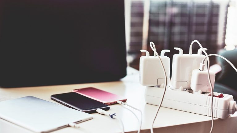 Energiesparen Auf einem Tisch liegen nebeneinander ein Tablet und zwei Smartphones. Все drei sind an eine Steckdosenleiste angeschlossen.