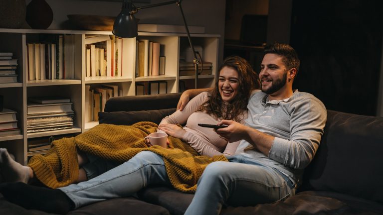 Laser-TV Ein Paar sitzt im abgedunkelten Wohnzimmer auf dem Sofa, eine Person hält eine Fernbedienung in der Hand.