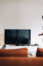 Zwei Personen vor einem Fernseher mit schwarzem Display, eine sitzt auf dem Sofa vor einem Laptop, die andere steht mit der Fernbedienung in der Hand.