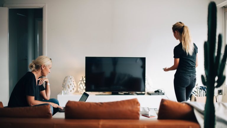 Zwei Personen vor einem Fernseher mit schwarzem Display, eine sitzt auf dem Sofa vor einem Laptop, die andere steht mit der Fernbedienung in der Hand.