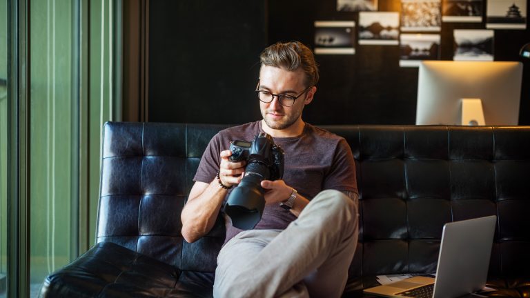 NAS einrichten Eine Person sitzt auf einem Sofa und betrachtet Fotos auf einer Spiegelreflexkamera. Daneben steht ein aufgeklapptes MacBook.