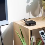 Ein Apple TV steht auf einem Regal neben einem Fernseher. Im Vordergrund hält eine Person eine Fernbedienung in der Hand.