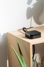 Ein Apple TV steht auf einem Regal neben einem Fernseher. Im Vordergrund hält eine Person eine Fernbedienung in der Hand.