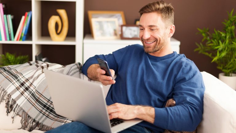 Google Authenticator einrichten Eine Person sitzt auf einem Sofa mit einem Laptop auf dem Schoß. In der Hand hält sie ein Smartphone.