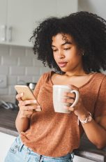 Eine Person steht mit einer Kaffeetasse in der Hand in der Küche und schaut auf ihr Smartphone.