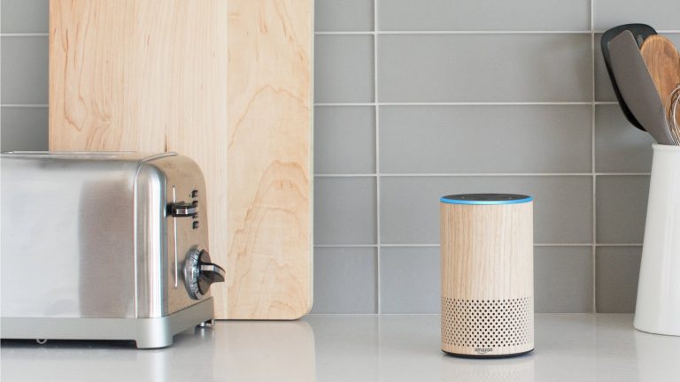 Alexa Sprachassistenz Ein Echo-Lautsprecher im Holz-Look steht auf einer Arbeitsplatte in der Küche neben einem Toaster und einigen anderen Küchenutensilien.