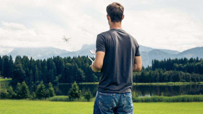 Drohne für Einsteiger Eine Person steht auf einer Wiese und lässt eine Drohne steigen, die am Horizont verschwommen über einer Baumreihe fliegt.