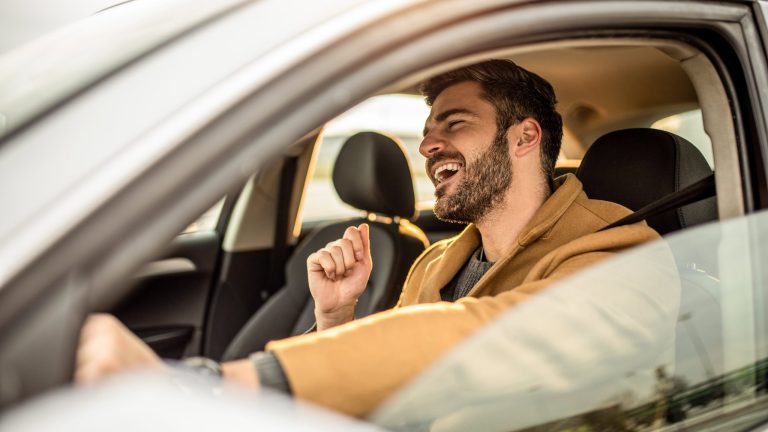 Bluetooth Lautsprecher Auto Eine Person sitzt in einem Auto, hat das Fenster auf der Fahrerseite heruntergelassen und singt.
