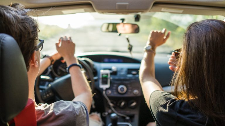 Zwei Personen sitzen in einem Auto und scheinen sich zu Musik zu bewegen. An der Mittelkonsole ist ein Smartphone angesteckt.