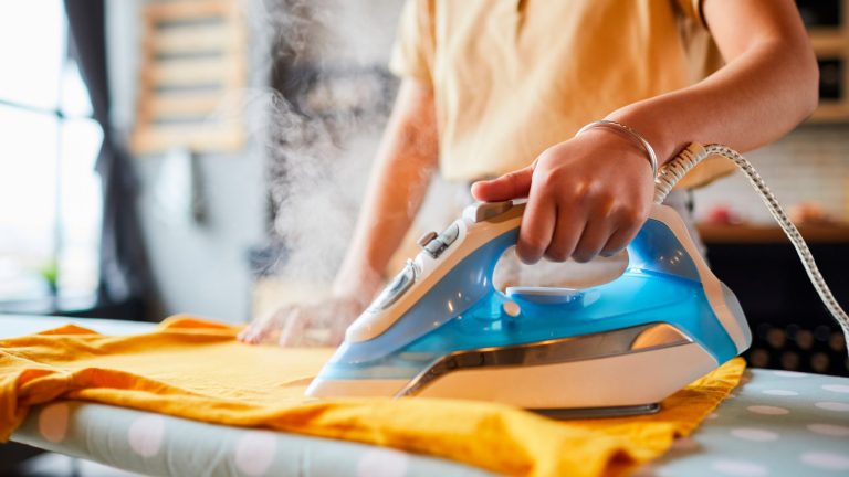 Bügeleisen entkalken Eine Person bügelt ein gelbfarbenes T-Shirt. Aus dem blautransparenten Bügeleisen entweicht Wasserdampf.
