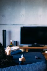 Eine Person sitzt auf einem Sofa vor einem Fernseher und hält eine Fernbedienung in der Hand.