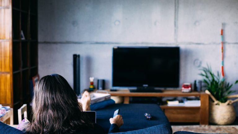 Eine Person sitzt auf einem Sofa vor einem Fernseher und hält eine Fernbedienung in der Hand.