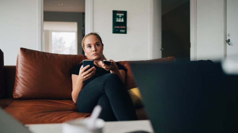 Fernseher reagiert nicht auf Fernbedienung Eine Person sitzt auf einem Sofa, in der Hand hält sie eine Fernbedienung und ein Smartphone.