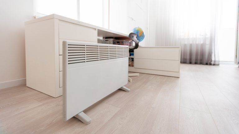 Eine Elektroheizung steht vor einem Schreibtisch am Boden.